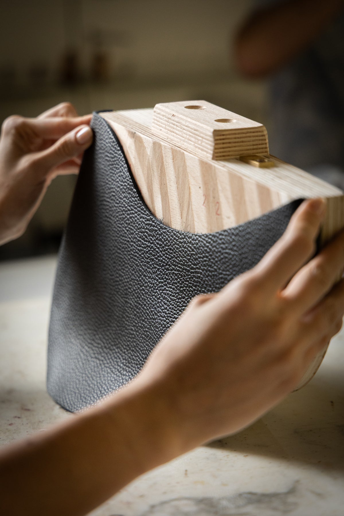 Hands holding a textured dark gray leather bag with a light wood handle and brass hardware.